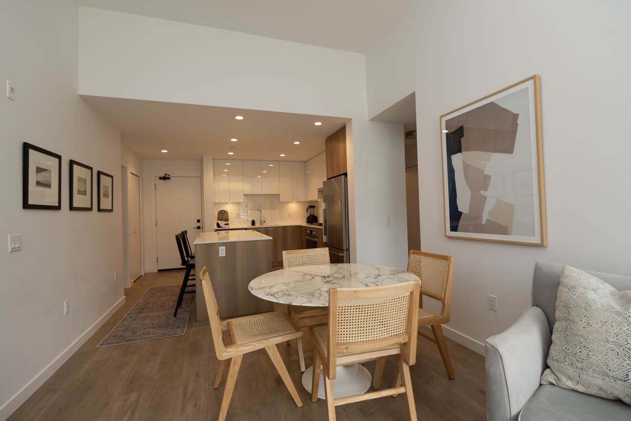 Furnished dining area with marble table and woven chairs, leading to a modern kitchen with white cabinets and stainless...