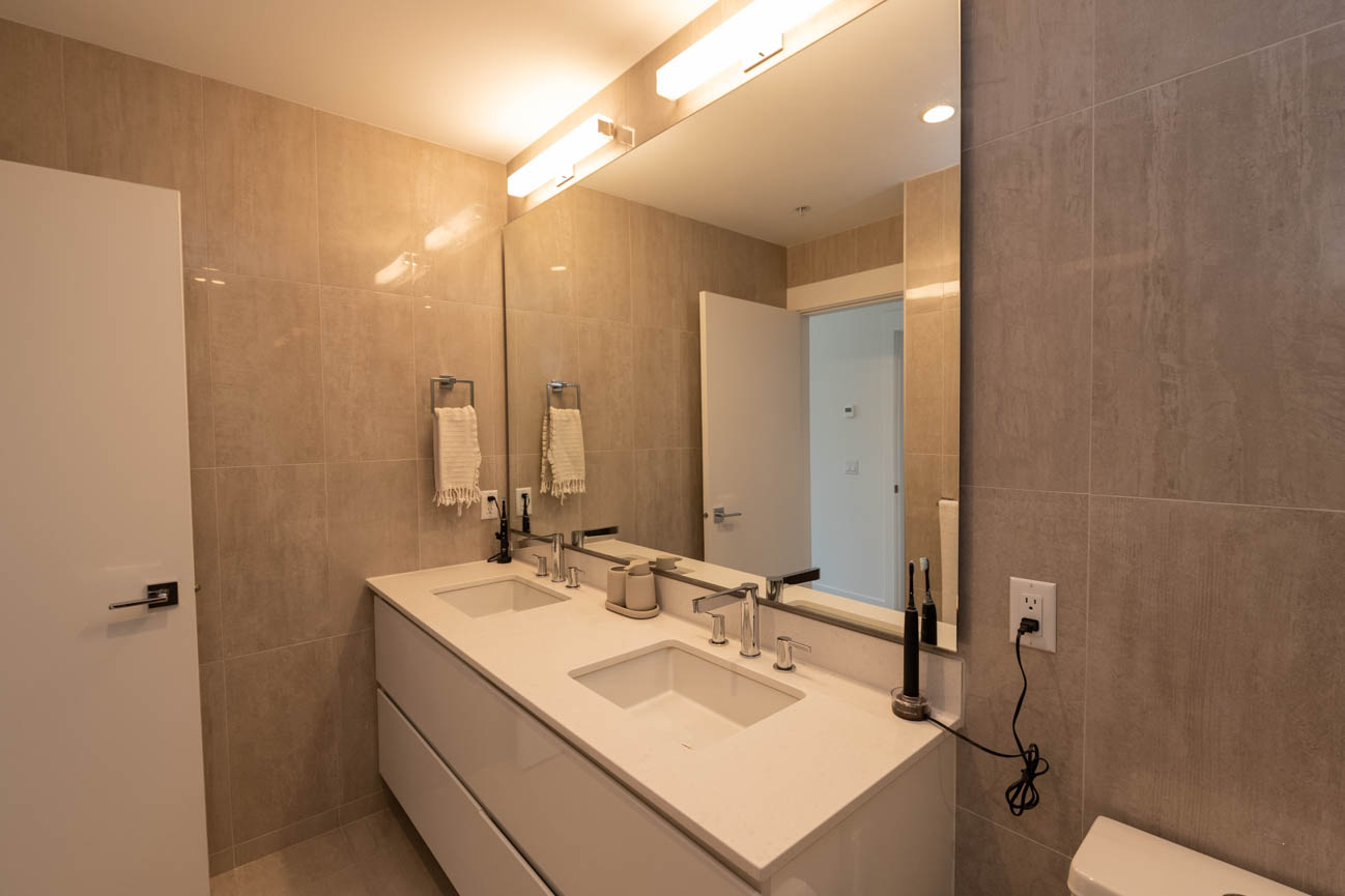 Furnished bathroom with double sink, modern lighting, and beige tile walls. - 308 - 715 W15th Street, North Vancouver