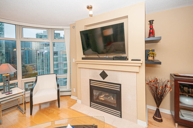 Spacious living room with a large window offering a city view, a fireplace, and a mounted TV. - 1605 - 1199 Marinaside Crescent, Vancouver Downtown