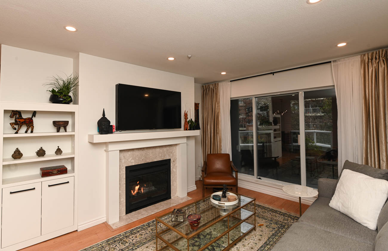 Furnished living room with hardwood floors, fireplace, and sliding glass door leading to balcony. - 208-128 8th Street West, North Vancouver