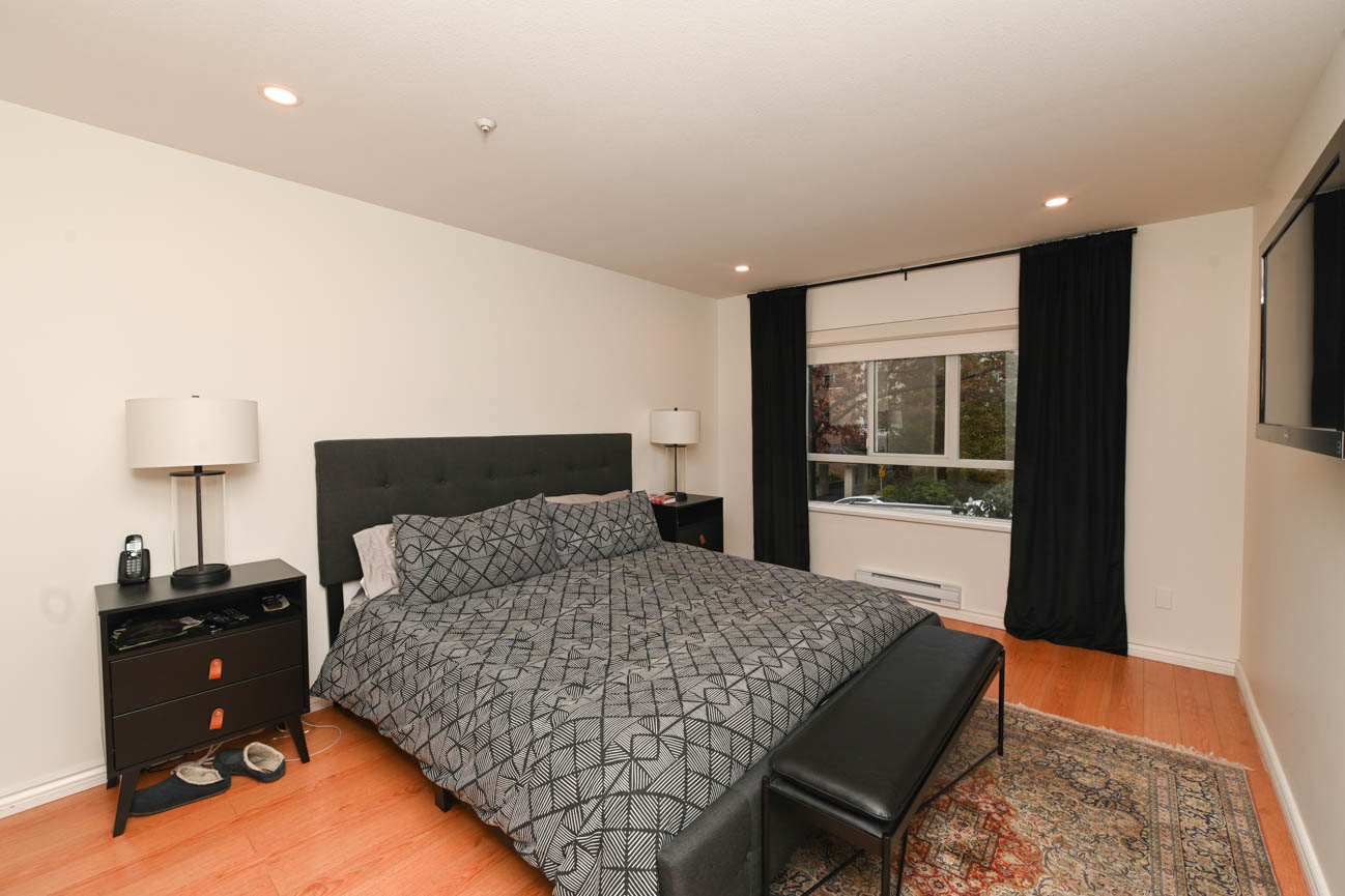 Furnished bedroom with hardwood floors, black curtains, and a geometric-patterned bedspread. - 208-128 8th Street West, North Vancouver