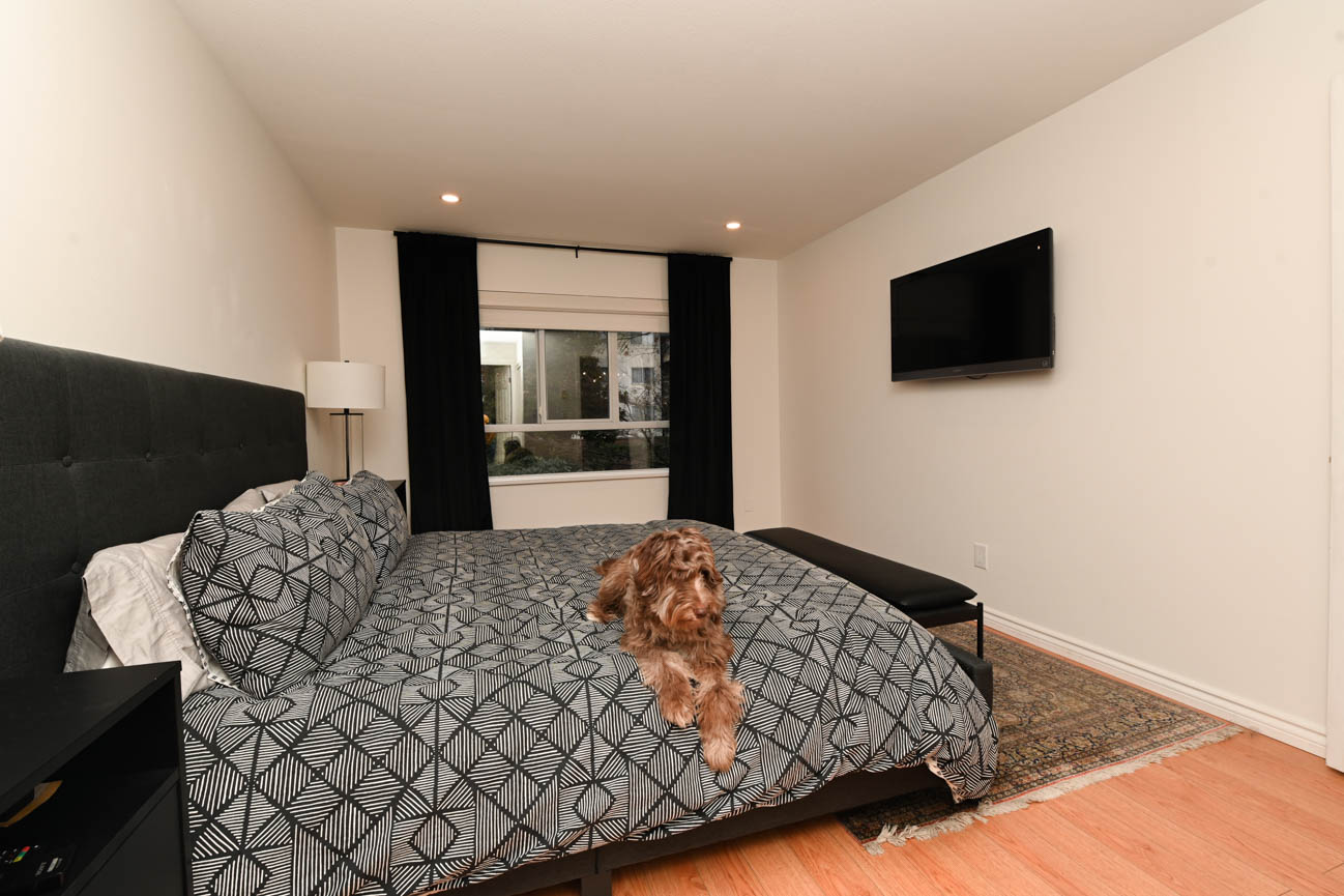 Furnished bedroom with hardwood floors and a cozy bed setup, featuring a mounted TV and a window with black curtains.