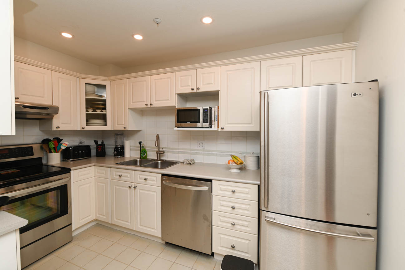 Bright kitchen with white cabinets, stainless steel appliances, and a bowl of fruit on the counter. - 208-128 8th Street West, North Vancouver