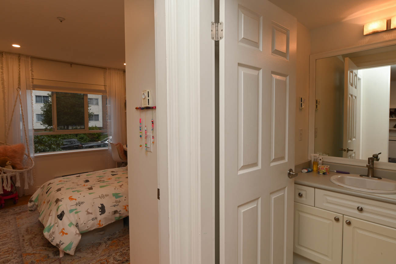 Bright bedroom with city view and cozy bedding, adjacent to a bathroom with white cabinets and a large mirror.