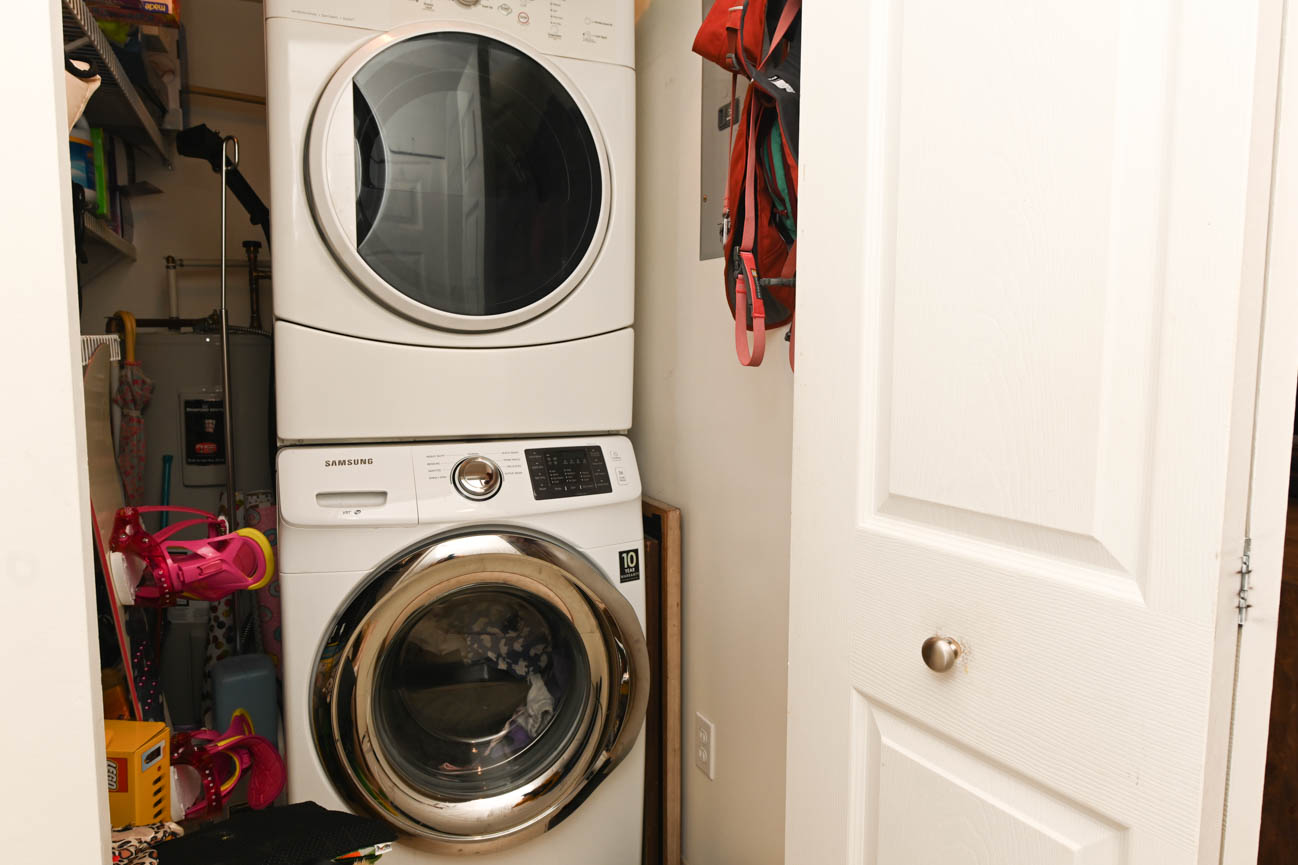Laundry room with stacked washer and dryer, cluttered shelves, and door leading to another space. - 208-128 8th Street West, North Vancouver