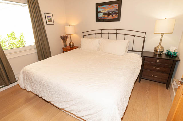 Spacious bedroom with white bedding, wooden floors, and a cityscape painting above the bed. - 2128 Grand Blvd, North Vancouver