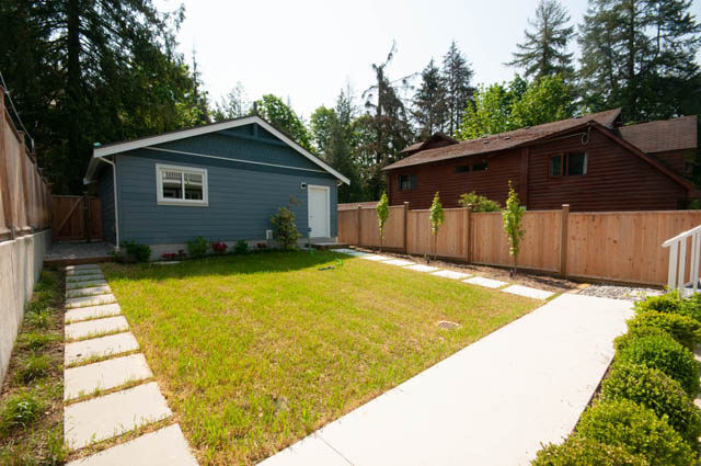 Spacious backyard with a modern blue house, wooden fence, and stepping stones leading to a red cabin. - Main - 1912 Larson Rd, North Vancouver
