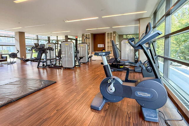 Gym space featuring Technogym stationary bike and rich hardwood flooring near expansive windows. - 504-3602 Aldercrest Drive, North Vancouver