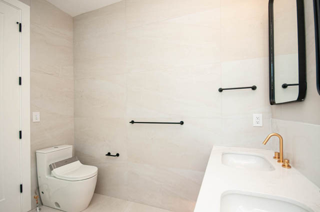 Modern bathroom with dual white sinks, gold fixtures, and black grab bars on beige tile walls. - 5909 Athlone Street, Vancouver West Side