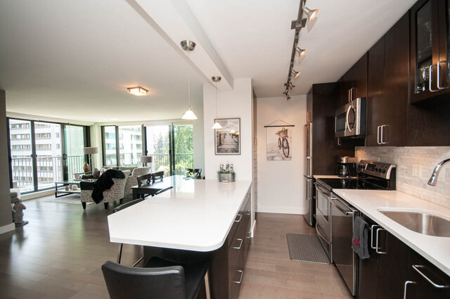 Open-concept kitchen with white countertops and dark cabinets, opening to a living room with hardwood floors and city views.