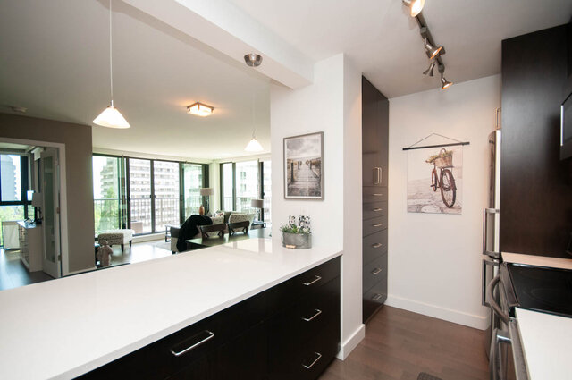 Modern kitchen with white countertops, dark cabinetry, and stainless appliances opening to a city-view living room.