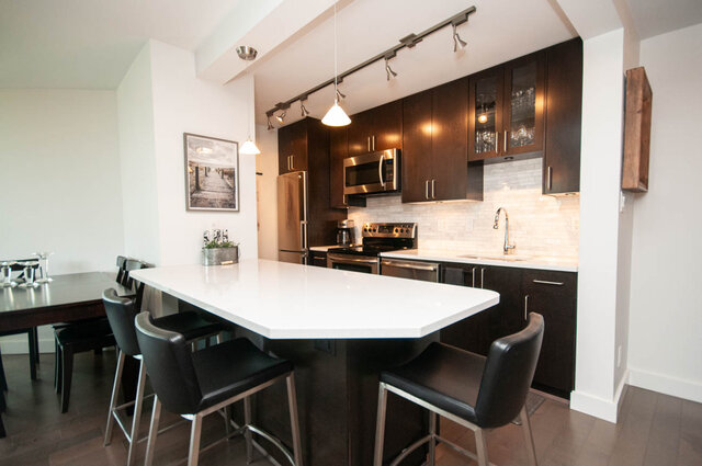 Modern kitchen with dark wood cabinets, white countertops, and a large island with black bar stools. - 1006-4165 Maywood Street, Burnaby