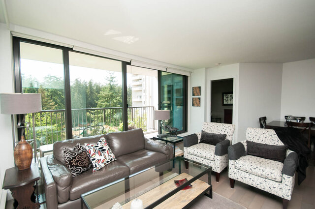 Furnished living room with brown leather sofa and glass coffee table, opening to a balcony with a green treetop view.