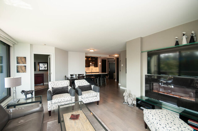 Furnished living room with patterned armchairs, hardwood floors, and a wall-mounted TV above a fireplace. - 1006-4165 Maywood Street, Burnaby