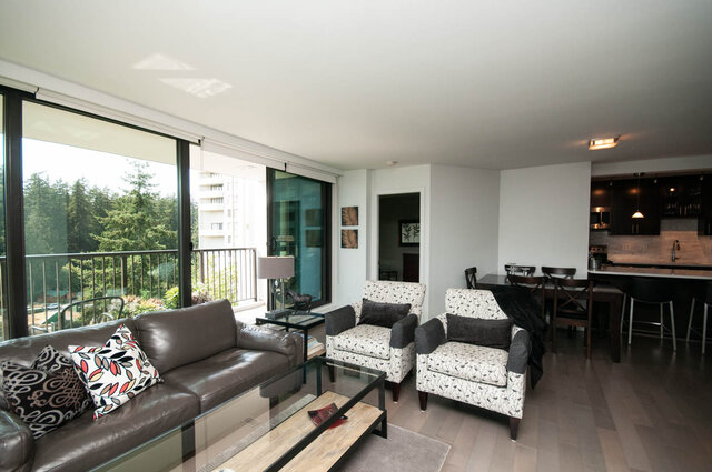 Furnished living room with brown leather sofa, patterned armchairs, and sliding doors to a balcony with tree views.