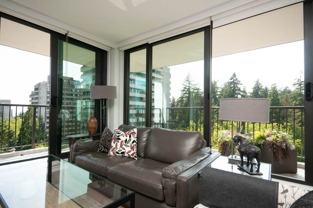Furnished living room with brown leather sofa and floor-to-ceiling windows offering a view of trees and high-rises.