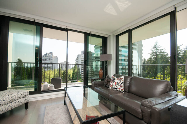 Furnished living room with a brown leather sofa and floor-to-ceiling windows framing a city and greenery view. - 1006-4165 Maywood Street, Burnaby