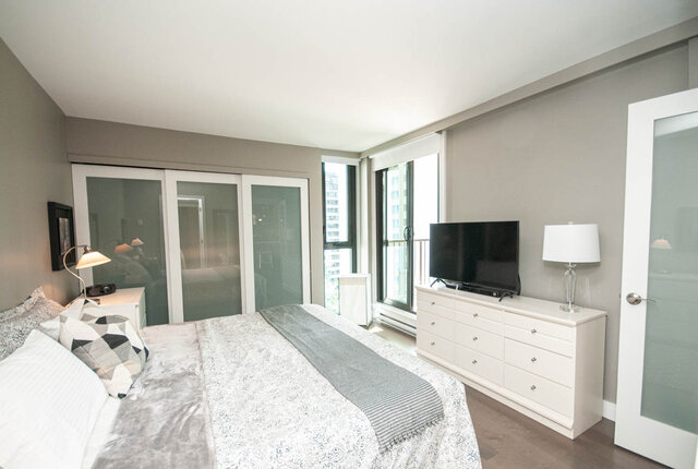 Furnished bedroom with a gray-dressed bed, white dresser with TV, and large windows offering a city view. - 1006-4165 Maywood Street, Burnaby