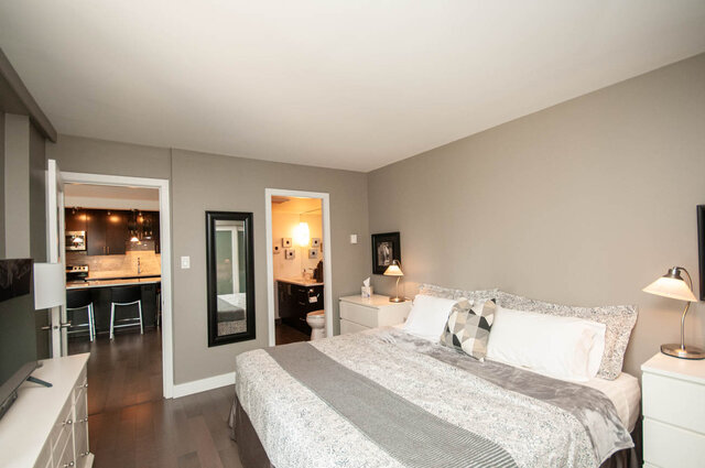 Furnished bedroom with gray bedding, hardwood floors, and open doorways to a dark-cabinet kitchen and bathroom. - 1006-4165 Maywood Street, Burnaby