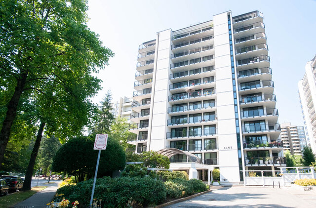 Modern white condo building exterior with stacked balconies and lush green tree landscaping. - 1006-4165 Maywood Street, Burnaby