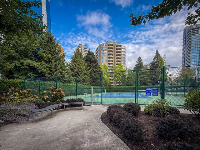 Landscaped courtyard with a curved bench overlooking a fenced tennis court and high-rise skyline. - 1006-4165 Maywood Street, Burnaby