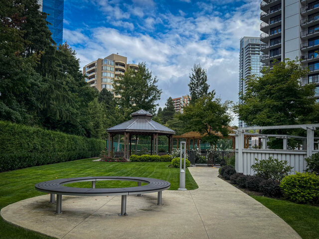 Lush outdoor courtyard with a circular bench, manicured lawn, and a wooden gazebo framed by high-rise towers. - 1006-4165 Maywood Street, Burnaby
