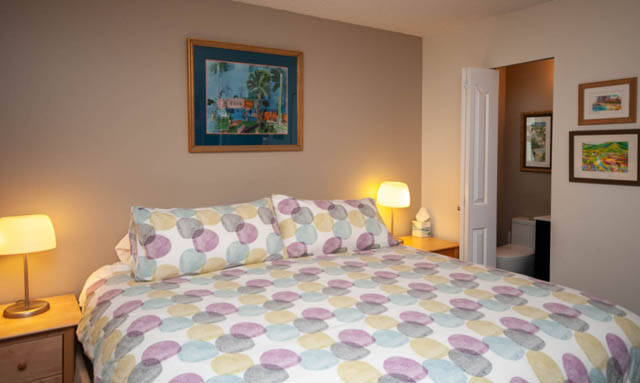 Bright bedroom with patterned bedding and bedside lamps, leading to another room. - 2077 W 13th Ave, Vancouver West Side