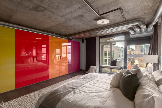 Modern bedroom with glossy multicolor sliding wardrobe panels and a city view through floor-to-ceiling windows.