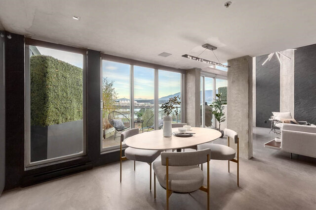 Dining area with a light wood table and upholstered chairs, framed by floor-to-ceiling windows with a water and mountain...