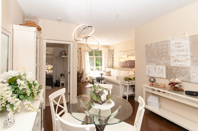 Dining area with a glass-top table, white chairs, and hardwood floors opening to a bright living room. - 102-1633 Mackay Avenue, North Vancouver