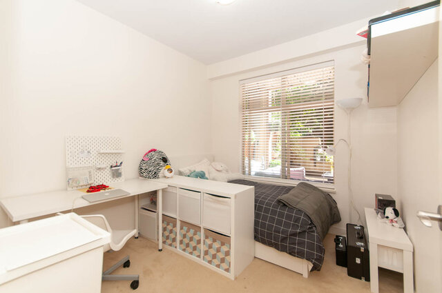 Bright bedroom with a white desk workspace, low bed, and storage bins beneath a window with blinds. - 102-1633 Mackay Avenue, North Vancouver