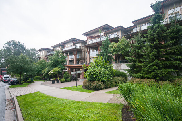Landscaped condo exterior with paved walkway, manicured lawn, and balconies framed by tall evergreen trees. - 102-1633 Mackay Avenue, North Vancouver