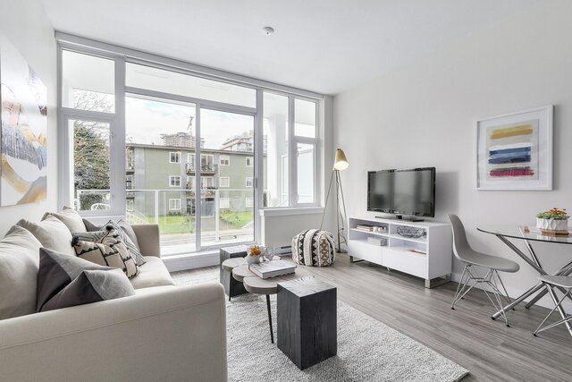 This is a living room with a sofa, coffee table, TV stand with television, large windows, and a dining area.