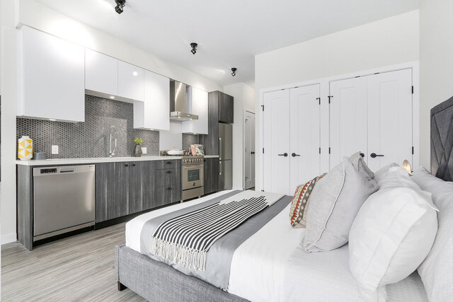 The bedroom has a bed with white bedding, a striped throw blanket, an integrated kitchen area with stainless steel appliances
