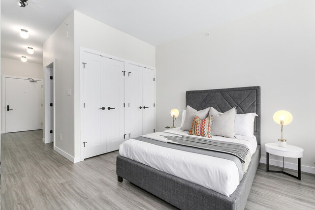 This bedroom contains a bed with white linens, two bedside lamps, built-in closet doors, and light wood flooring.