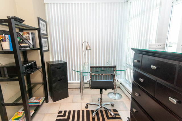Bright home office with glass desk, black rolling chair, and vertical blinds, showcasing a city view.
