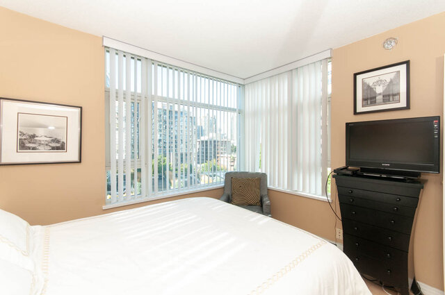 Spacious bedroom with large windows offering a city view, featuring a neatly made bed and a dresser with a TV above.