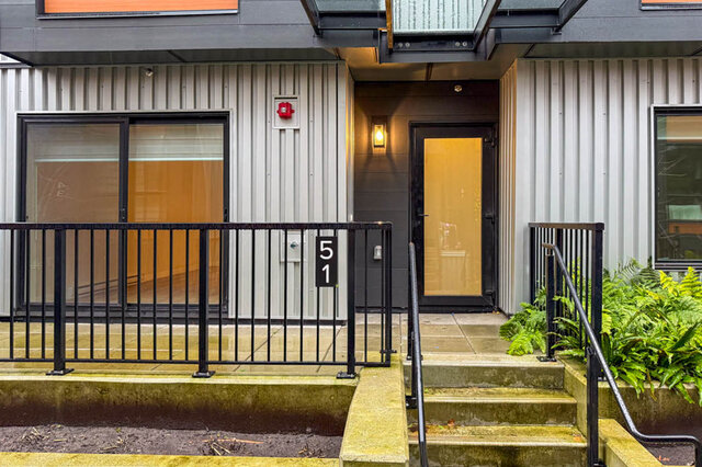 The entrance features a black metal railing, numbered address plaque, concrete steps, and greenery beside the steps.