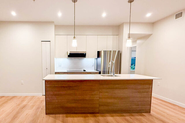 The kitchen features a central island with sink, stainless steel refrigerator, white cabinets, hardwood floors, and pendant l
