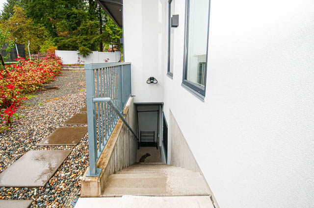 Furnished patio with stone path leading to white wall and door, surrounded by greenery. - Suite 2 - 788 Montroyal Boulevard, North Vancouver