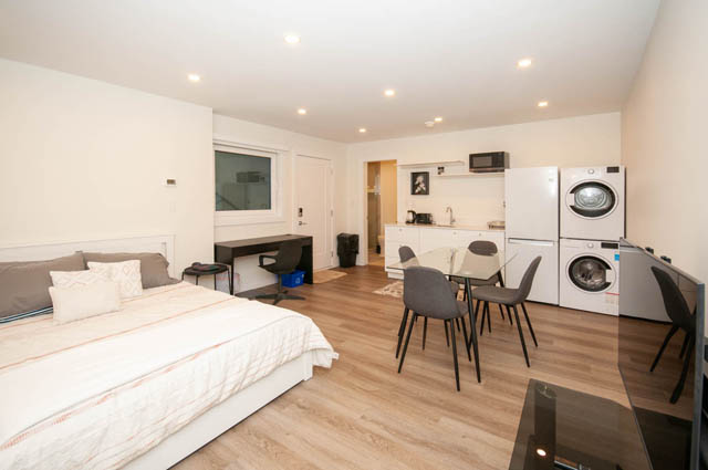 Bright bedroom with white bedding and wooden floors, adjacent to a compact kitchen with white countertops and a washer/dryer.