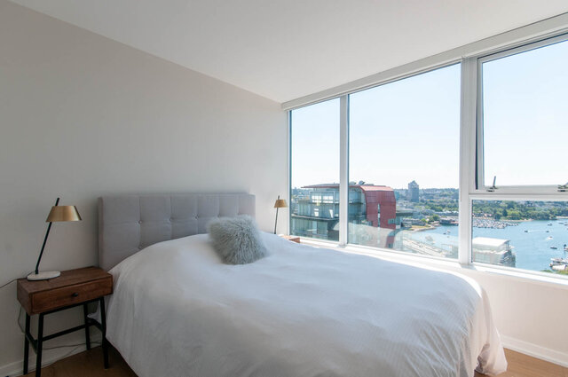 Spacious 1-bedroom + den condo with a serene city view and large windows, featuring a cozy bed setup and modern decor.