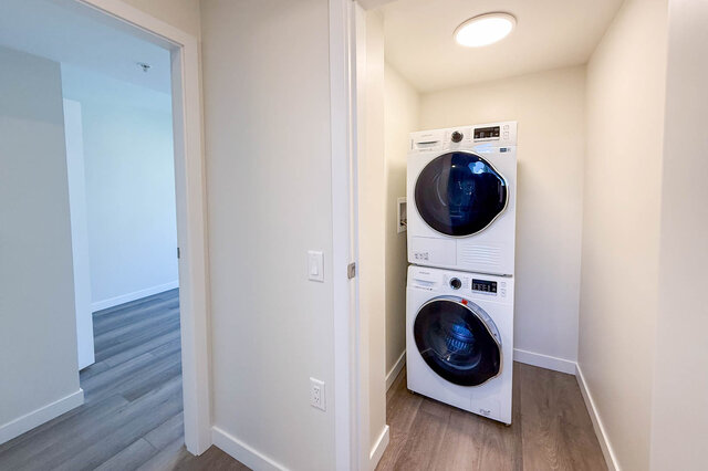 Bright laundry room with stacked washer and dryer, hardwood floors, and a door leading to another room.