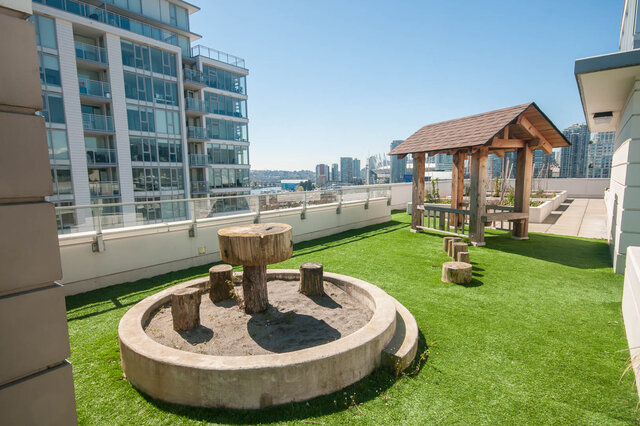 The balcony features artificial turf, a wooden shelter with a roof, tree stump seating, and a city view beyond the railing.