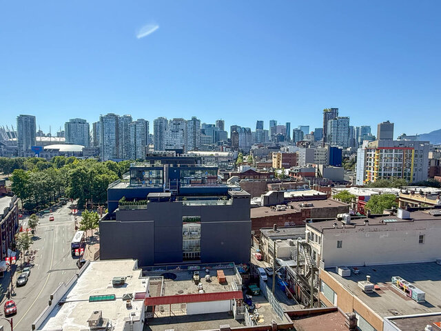 The view shows a city skyline with tall buildings, green trees, a street with vehicles, and a clear blue sky.