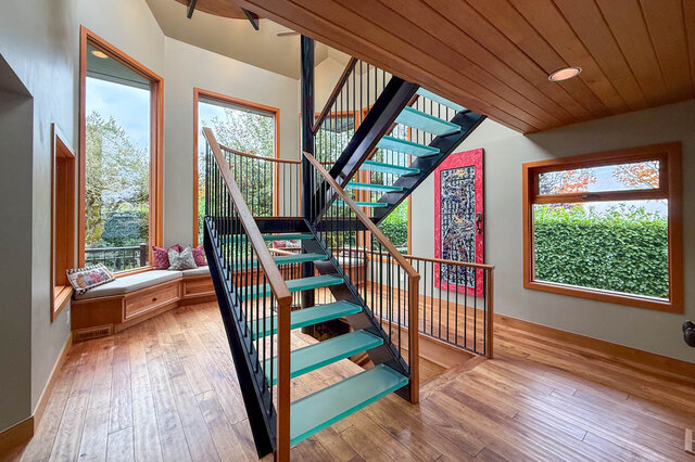 Staircase with teal glass steps and wooden railings leads to upper level, featuring large windows with a view of greenery.