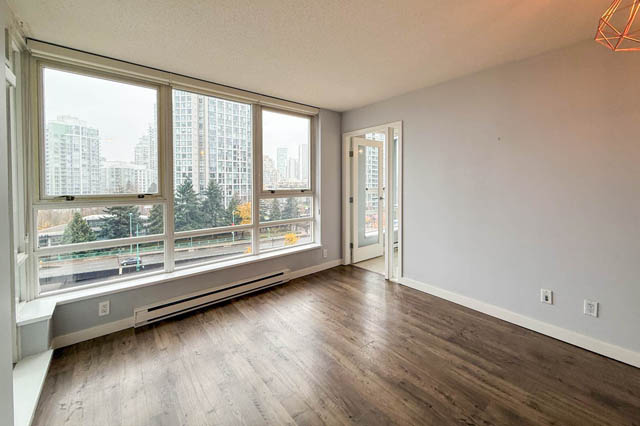 Spacious living room with hardwood floors, large windows offering a city view, and a door leading to another room.