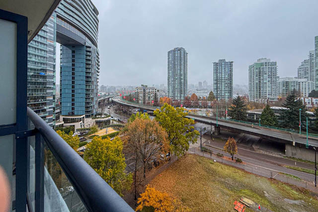 Furnished 1-bedroom + den with stunning city view and modern balcony overlooking a bustling urban landscape.
