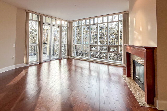 Spacious living room with hardwood floors, large windows, and a fireplace. - 301-605 Clyde Ave, West Vancouver