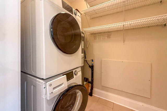 Compact laundry room with stacked washer and dryer, white shelving, and tiled floor. - 301-605 Clyde Ave, West Vancouver